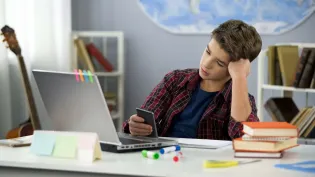 A young boy sitting at a desk with his head resting on his fist. He is looking at his mobile phone. He has a laptop with sticky-notes and is surrounded by clutter. He is struggling to focus.