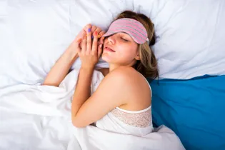 A college age female sleeping in her bed. She is wearing a pink and grey striped face mask. Her duvet cover and pillows are white with a blue sheet. Her nails are painted purple, red, pink, yellow, and orange.