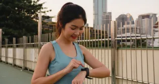 Young woman with black hair smiling. She is wearing a teal running shirt and is tapping her black sports watch. She is running with a blurred city background.