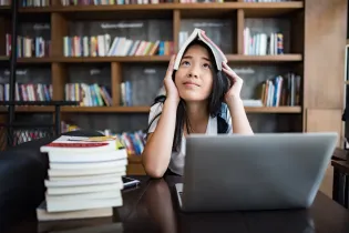 Young woman with black hair sitting at her desk with her laptop. She has a stack of textbooks next to her and is holding one on top of her head. She looks demotivated.