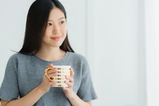 Young woman with black hair smiling. She is wearing a dark grey t-shirt and is holding a bronze and white striped mug.