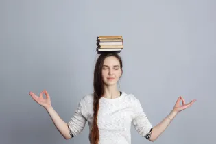 A young female with a long dark-brown ponytail sitting with her arms out and elbows bent at 45 degrees. She has 5 books balanced on her head.