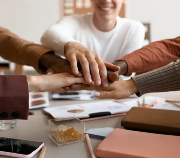 A group of students collaborating on a project, with their hands joined in the center of a table, symbolizing teamwork and support.