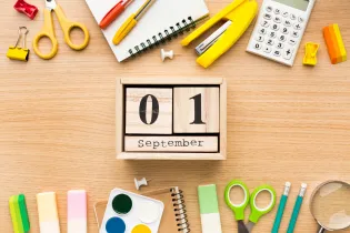 A wooden desk with blocks of wood on it saying, '01 September'. Surrounding the center piece are some yellow and green scissors, a yellow stapler, a white calculator, a yellow sharpener, and some pencils and erasers.