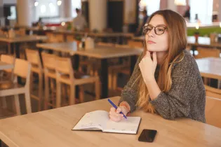 Young female with light brown hair. She is holding a blue pen and is writing notes in her diary. She is wearing a brownish-grey jersey and is sitting in a coffee shop. The background has blurred wooden desks and chairs.