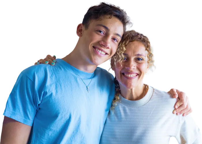 Male boy with brown curly hair smiling. He is standing shoulder to shoulder with his mom next to him. She has blonde curly hair.
