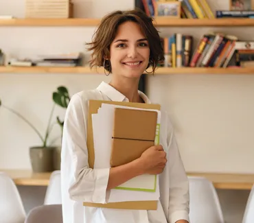 A tutor smiling and holding a stack of books in a tutoring center, with shelves of books in the background.