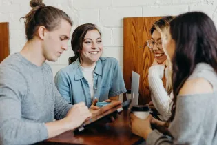 A group of 4 college-age students sitting at a desk smiling and talking to each other. There are three females and one male.