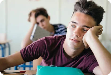 Two boys in class, half asleep with their heads resting on their hands. Their eyes are droopy and they might fall asleep any second.