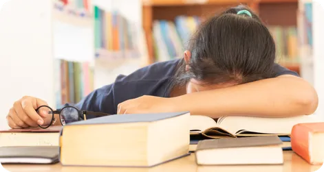 Young brunette student feeling defeated. She is lying with her head on her desk, surrounded by books.
